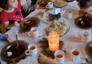 Dzieci siedzą przy stole nakrytym białym obrusem. Na środku stołu talerz z kapustą z grochem oraz świecznik i gałązki igliwia.